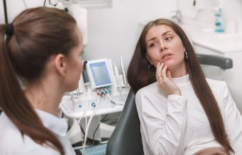 A woman in a dental chair touches her cheek while talking to a dentist in a clinic setting.